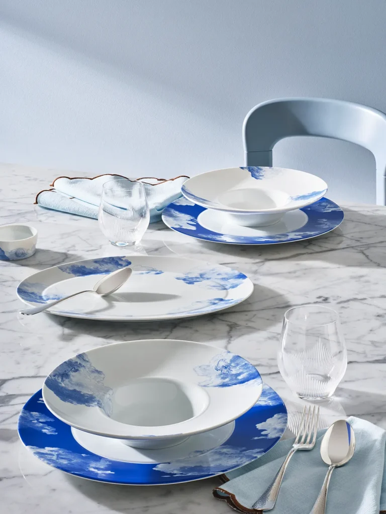 Fürstenberg RÊVERIE-Porzellan-Suppenteller mit zarten blauen Wolkenmotiven, die auf einem Marmortisch mit pastellfarbenen Servietten, elegantem Besteck und geschwungenen Gläsern angerichtet sind.