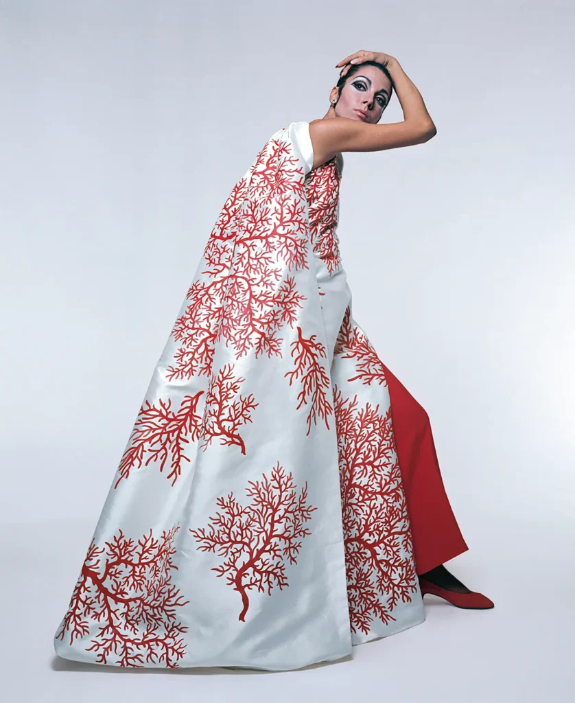 Ritratto in studio di una modella con abito bianco Valentino ricamato con corallo rosso, pantaloni rossi e trucco marcato.